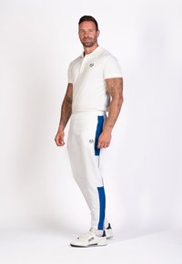 Polo shirt blanc à manches courtes, pantalons de jogging blancs avec des accents bleus, et baskets blanches avec des détails navy ; un homme se tient avec confiance.