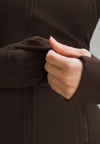 Brown sports jacket with a zipper, featuring form-fitting sleeves and thumbholes. Smooth, stretchy fabric with contrasting stitch details.