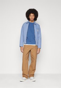Chaqueta de denim azul claro con un bolsillo en el pecho, suéter azul de cuello redondo, pantalones de trabajo beige y zapatillas de lona grises. Texturas suaves en todo.