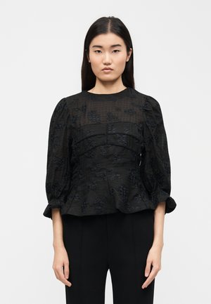 Kobieta o długich ciemnych włosach, ubrana w czarną teksturowaną bluzkę z bufkami na rękawach i czarne spodnie, stojąca na jednolitym tle.