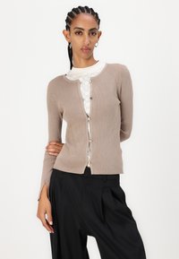 Beige ribbed cardigan with a round neckline, front zipper, and side slits. Worn over a patterned white top. Black wide-leg trousers.