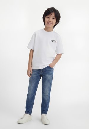 Χαμογελαστός αγόρι με μαύρα μαλλιά στέκεται φορώντας λευκό T-shirt DSQUARED2, μπλε τζιν και λευκά αθλητικά παπούτσια σε ουδέτερο λευκό φόντο.