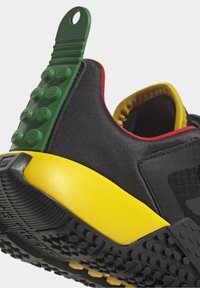 Svart träningsskor med en strukturerad yta, grön gummiklack, gul mellansula och svart grepp-sula. Röda och gula detaljer synliga.