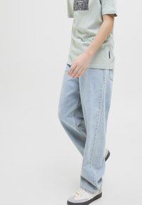 Ljusblå jeans med vida ben och en avslappnad passform. Kombinerat med en grön t-shirt med ett mönstrat motiv på framsidan och vita sneakers.