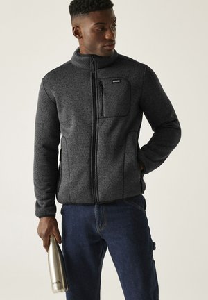 Hombre con chaqueta polar gris oscuro con cremallera y vaqueros azules, sosteniendo una botella de agua de acero inoxidable con la mano izquierda.