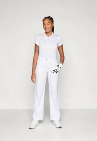 Mulher em pé, sorrindo, vestindo uma polo branca, calças brancas e ténis brancos, segurando uma luva de golfe branca com detalhes pretos na mão direita.