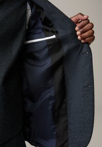 Interno di una giacca blu navy con fodera in raso scuro, una tasca con bordino bianco e dettagli di cucitura visibili. Mano che stringe il risvolto.