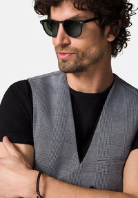 Kamizelka w czarno-białe wzory ma teksturowany wzór w kratkę, zestawiona z czarną koszulką. Osoba nosi ciemne okulary przeciwsłoneczne.