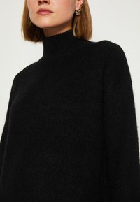Maglione nero a collo alto realizzato in un materiale morbido e texturizzato. Vestibilità ampia con spalle scese. Design minimalista, senza motivi o dettagli visibili.