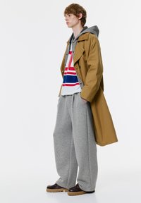 Bežan plašč s črtastim kapuco in grafičnim majico. Sivi široki trenirki in temni mokasini z debelim podplatom dopolnjujejo videz.