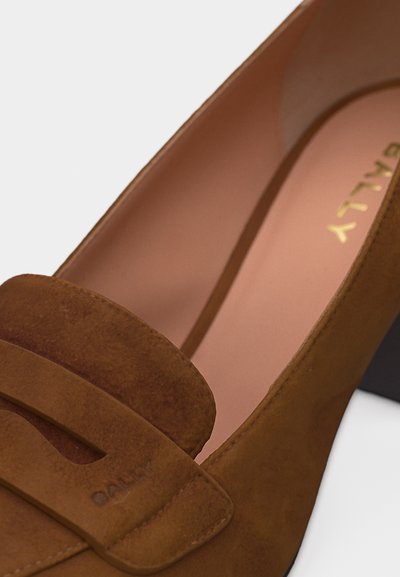 Mocassin en daim marron avec un petit talon carré, doté d'une lanière sur le devant et d'une doublure intérieure en cuir rose doux.