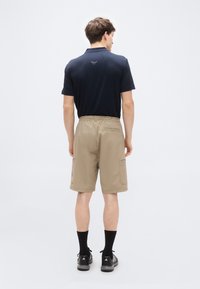 Μπλε σκούρο μπλουζάκι  πόλο με κοντά μανίκια, μπεζ cargo βερμούδα με ελαστική μέση, πλαϊνές τσέπες και μαύρα παπούτσια με σκούρες κάλτσες. Μινιμαλιστικός σχεδιασμός.