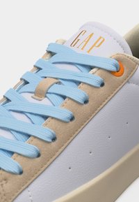 Baskets blanches avec des accents en daim beige, des lacets bleu clair, un œillet orange, et le logo "GAP" en jaune sur la languette.