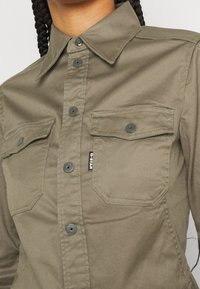 Olivgrön skjorta med knäppning som har krage, två bröstfickor och grå knappar. Tillverkad av ett texturerat, hållbart tyg med synliga sömmar.