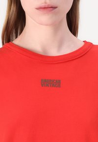 Czerwona bawełniana koszulka z okrągłym dekoltem i krótkimi rękawami, z szarym logo "AMERICAN VINTAGE" na klatce piersiowej. Gładka tekstura.