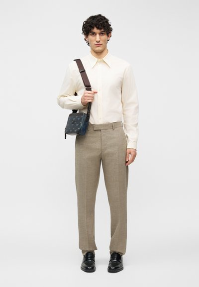 Άνδρας που φοράει κρεμ πουκάμισο με κουμπιά, μπεζ παντελόνι, μαύρα παπούτσια, κρατώντας μπλε τσαντάκι χιαστί με σκουρόχρωμο λουρί σε λευκό φόντο.