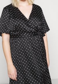 Schwarzes Satin-Wickelkleid mit kurzen Ärmeln, das weiße Polka-Dots, einen tiefen V-Ausschnitt und ein gebundenes Taillendetail aufweist. Glatte Textur.