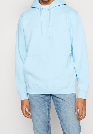 Mand iført lyseblå hættetrøje med japansk tekst på brystet og blå jeans, stående mod en ensfarvet baggrund.