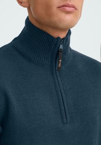 Dunkelblauer gestrickter Pullover mit hohem Kragen, ausgestattet mit einem halben Reißverschluss und gerippter Textur. Branding sichtbar am Reißverschluss-Anhänger.