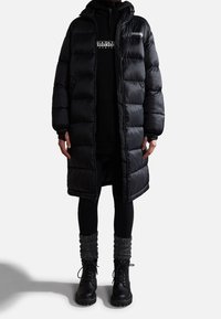 Abrigo puffer negro con capucha, con un acabado brillante, cierre de cremallera y puños acanalados. Lleva puesto un suéter negro y leggings con calcetines grises.