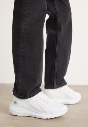 Baskets blanches Lacoste portées avec un jean noir, debout sur un sol beige devant un fond uni.