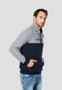 Sudadera con capucha azul marino y gris con textura a rayas, bolsillo frontal y capucha con cordón. Presenta detalles de logo y texto en las mangas.