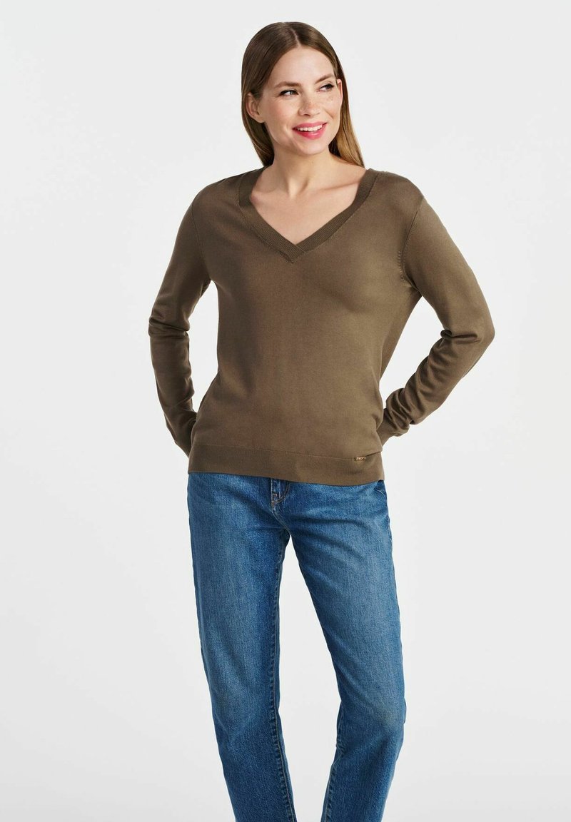 Zielony oliwkowy sweter w serek z miękkiego materiału, z długimi rękawami i prążkowanymi akcentami przy dekolcie i hemie. Połączony z niebieskimi dżinsami.