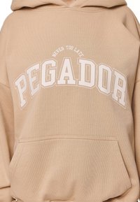 Sudadera beige de tejido suave con un bolsillo tipo canguro, un gran gráfico "PEGADOR" y el texto impreso "NUNCA ES TARDE" arriba.