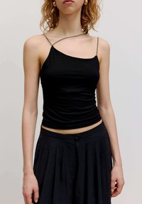 Schwarzes, tailliertes Tanktop mit einem Träger, aus glattem Stoff. Kombiniert mit hochgeschnittenen, gefalteten schwarzen Hosen.