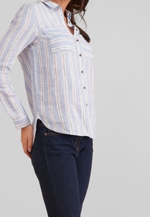 Femme portant une chemise à rayures bleu clair et blanche avec boutons, deux poches poitrine, et un jean bleu foncé, debout avec une main sur la hanche.