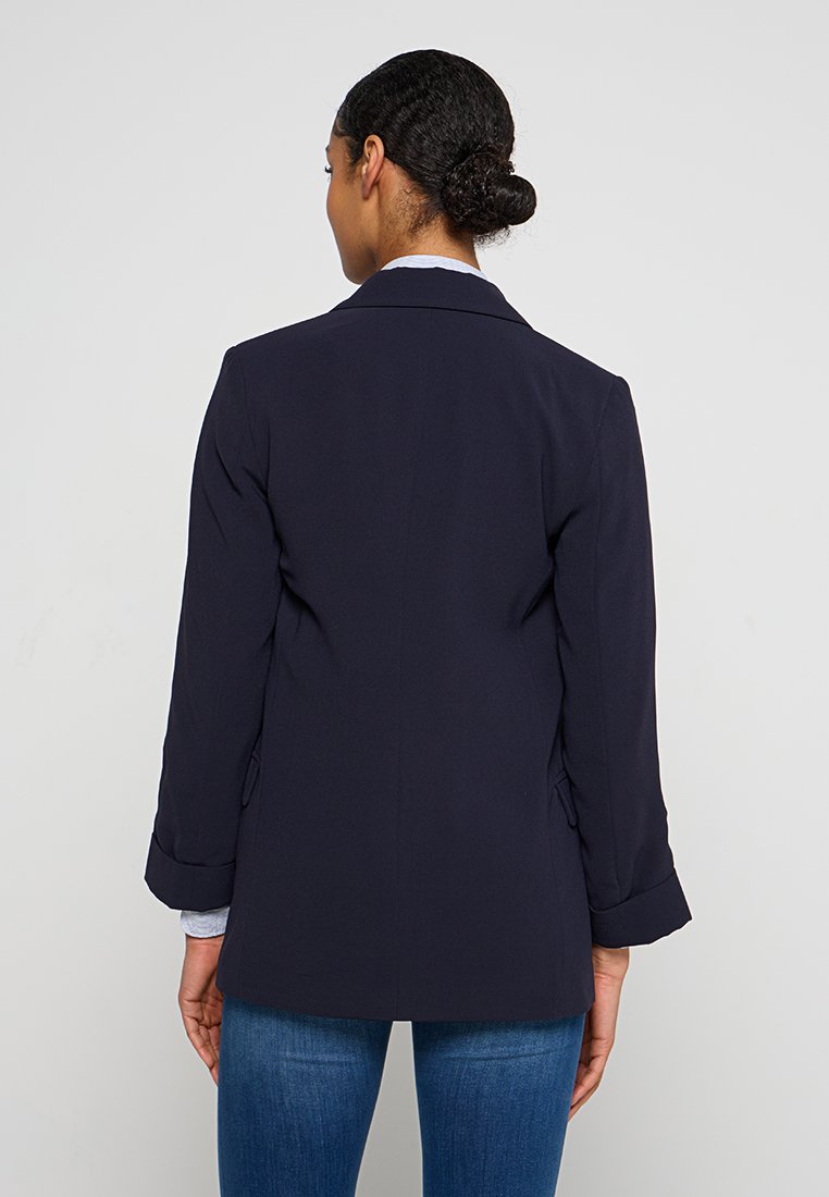 Donna con capelli scuri raccolti in un chignon, che indossa un blazer blu navale e jeans blu, vista da dietro su uno sfondo chiaro e uniforme.