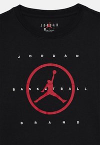 Melna kokvilnas T-krekls ar lielu sarkanu basketbola logo, kurā ir siluets un teksts "JORDAN BASKETBALL BRAND" baltā krāsā.