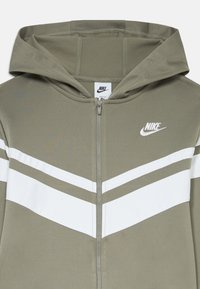 Olīvu zaļa jaku ar rāvējslēdzēju un baltu ševronu rakstu, kas ir aprīkota ar kapuci. Satur priekšējo rāvējslēdzēju un Nike logo uz krūšu daļas. Gluda auduma tekstūra.