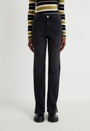 Personne portant un jean droit noir avec des fentes latérales, des chaussures noires épaisses et un haut à manches longues rayé noir, blanc, beige et vert.