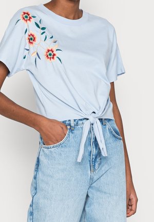 Vaaleansininen lyhythihainen toppi, jossa etuosassa solmuke ja punainen kukkaommel olkapäällä, yhdistetty vaaleansinisiin korkeavyötäröisiin farkkuihin.