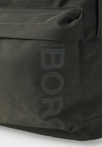 Mochila de tecido verde escuro com um grande logotipo "BORG" em cinza, material texturizado, com costura de contraste e um bolso lateral.