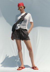 Jeune personne portant un bandana rouge, un t-shirt graphique blanc, un short noir, un sac bandoulière noir et des tongs rouges sur un fond clair uni.