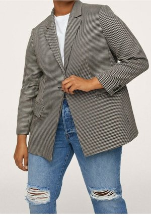 Osoba ubrana w czarno-białą marynarkę w jodełkę na białej koszuli i podniszczonych niebieskich dżinsach, zapinająca guzik marynarki.