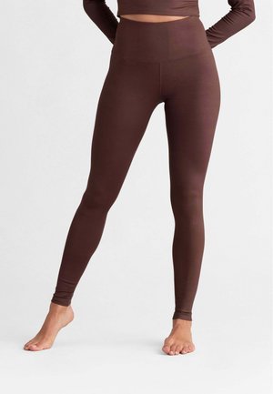 Frau trägt hoch taillierte dunkelbraune Leggings und passendes langärmeliges Crop-Top, steht barfuß mit den Händen in der Hüfte.