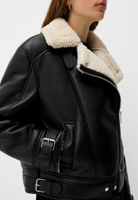 Giubbotto di pelle nera con colletto in finta pelliccia crema, distintivi accessori in argento e cinturini regolabili con fibbie sui lati.