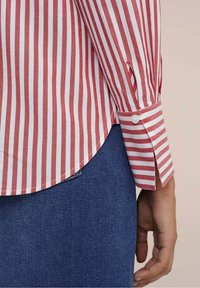 Red and white striped shirt with long sleeves, buttoned cuffs, curved hem, and soft cotton fabric, paired with dark denim.