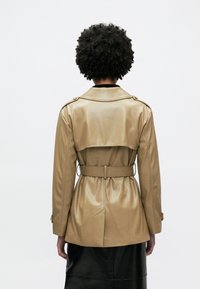 Ταν χαρακτηριστικό πανωφόρι από συνθετικό δέρμα με ζώνη στη μέση, επιθεωρήσεις στους ώμους και διπλό κούμπωμα. Ομαλή υφή, μεσαίου μήκους.