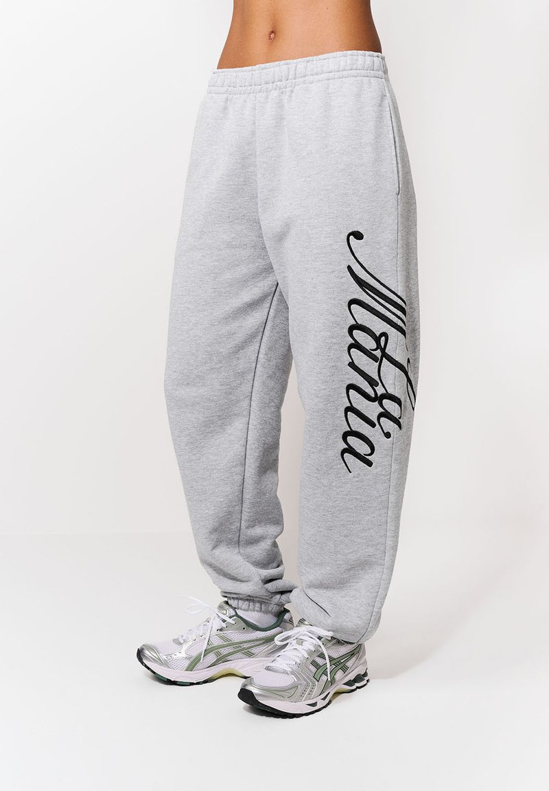 Osoba oblečená v svetlosivých teplákoch s čiernym nápisom na ľavej nohe a v bielych bežeckých topánkach so zelenými akcentmi, stojaca na bielom pozadí.