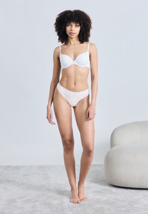 Femme aux cheveux bouclés debout pieds nus sur un tapis, portant un soutien-gorge et une culotte blancs, à côté d'un fauteuil blanc moderne arrondi.