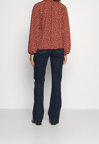 Persona che indossa jeans svasati blu scuro e una blusa floreale rossa a maniche lunghe, in piedi contro uno sfondo bianco uniforme.