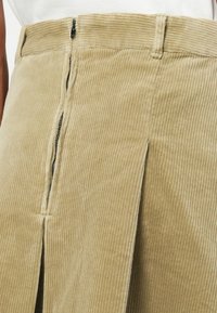 Gonna in corduroy khaki con zip frontale, righe verticali e dettaglio con spacco laterale. Il tessuto ha una texture morbida e un design strutturato.