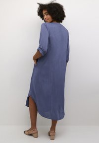 Robe à manches longues, longueur genoux en tissu bleu atténué. Présente des manches retroussées, des poches latérales et un ourlet légèrement asymétrique.