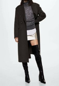 Long manteau vert foncé à double boutonnage, superposé à un col roulé gris et une chemise blanche, associé à une jupe argentée et des bottes noires montantes.