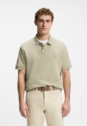 Young man with short curly hair wearing a beige polo shirt with a small embroidered logo and cream-colored pants with a brown belt.