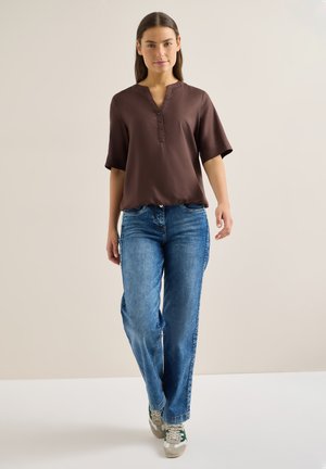 Kobieta idąca do przodu, ubraną w brązową bluzkę z krótkim rękawem, niebieskie dżinsy i szare trampki z zielonymi akcentami na jednolitym beżowym tle.